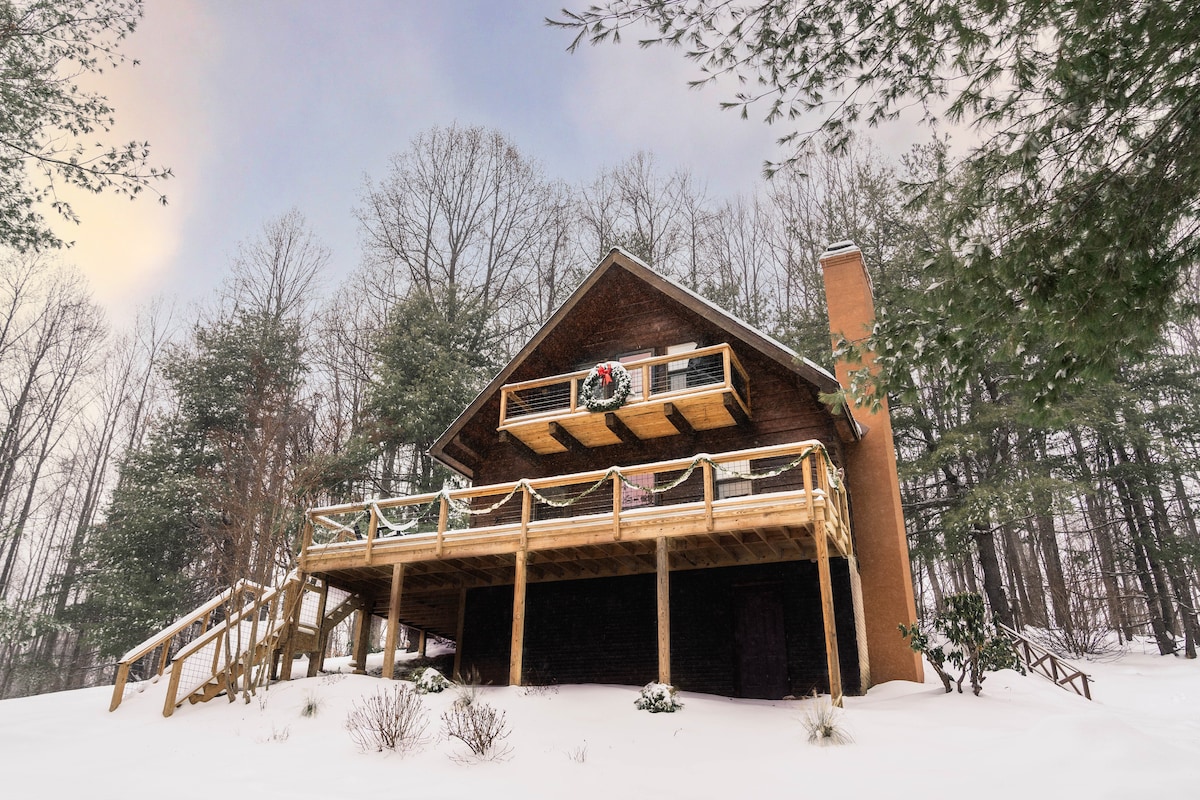 The cabin is set amidst a winter landscape, with a snow-covered ground and trees surrounding it. A wooden wrap-around deck features a festive wreath on the front, offering a welcoming appearance. The structure's angled roof and large windows enhance its rustic charm.