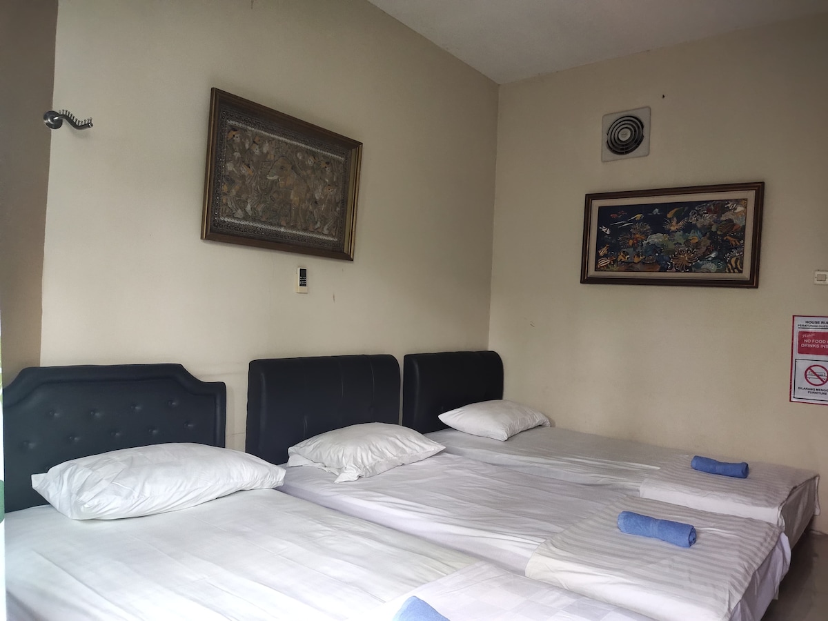 A spacious room features two single beds with white linens and contrasting headboards. Soft, rolled towels are placed on the beds. Wall art enhances the ambiance, and an exhaust fan is mounted above, ensuring a comfortable environment.