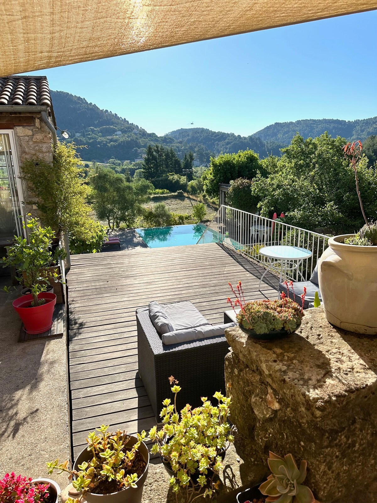 A sunlit terrace overlooks a landscaped garden and a salt-water pool, set against a backdrop of rolling hills. Comfortable seating is available, surrounded by vibrant greenery and flower pots, creating a serene outdoor space that invites relaxation.