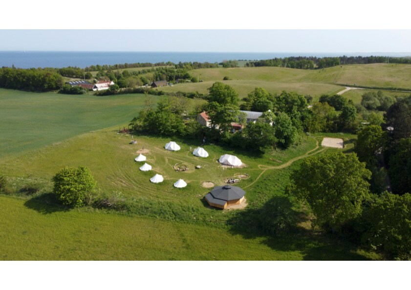 Aerial view of a glamping site featuring multiple white tents set amid lush greenery. Surrounding fields are visible, and the coastline lies in the distance. A communal area with a circular structure is located to the right, inviting gatherings in nature.
