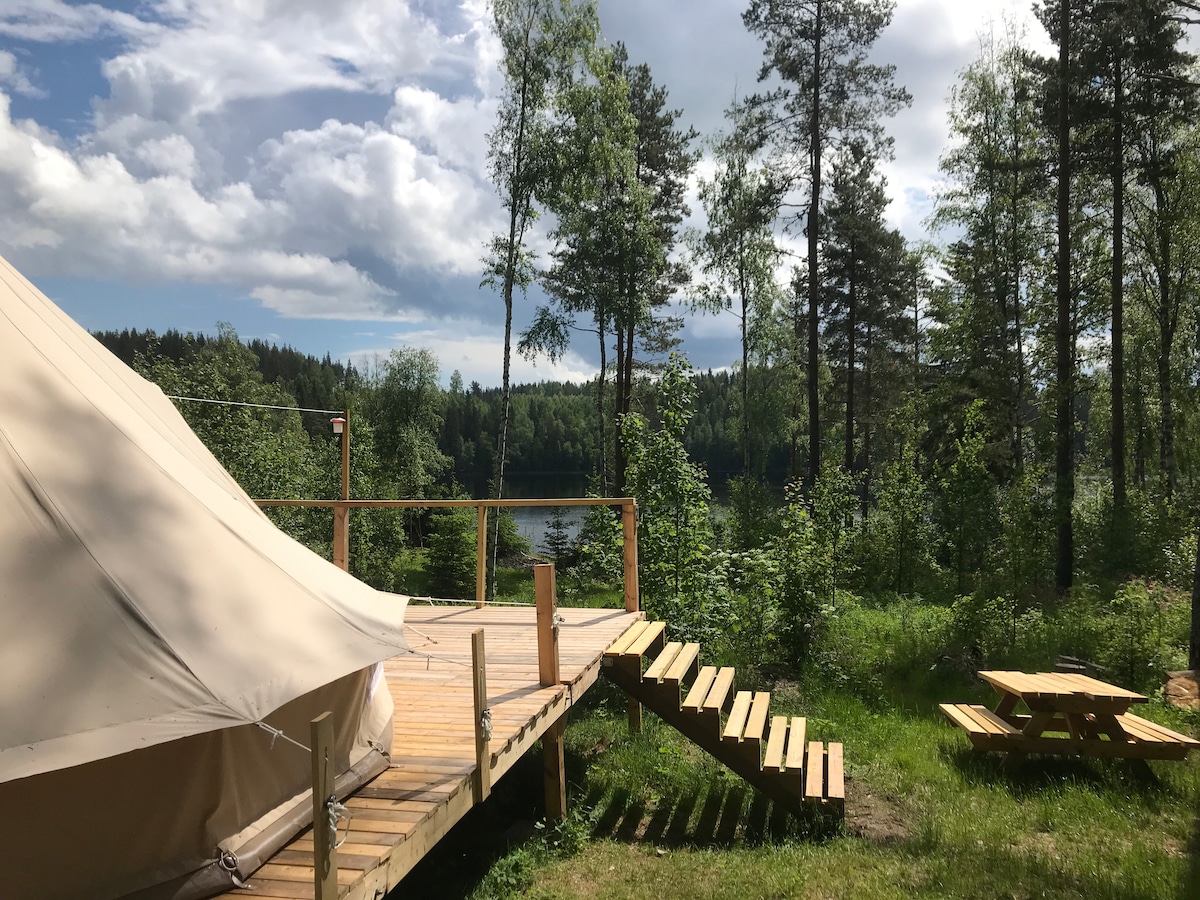A large tent is positioned on a wooden platform with a staircase leading down to a grassy area. Lush green trees surround the space, and a tranquil lake is visible in the distance under a partly cloudy sky.