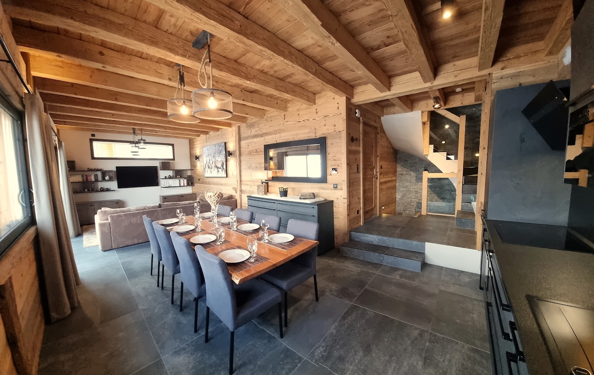The open-plan living area features a rustic wooden ceiling and walls, with a dining table set for six. A cozy sofa and entertainment unit are positioned nearby, while a staircase leads to the upper levels. Earthy tones and stone flooring contribute to a warm atmosphere.