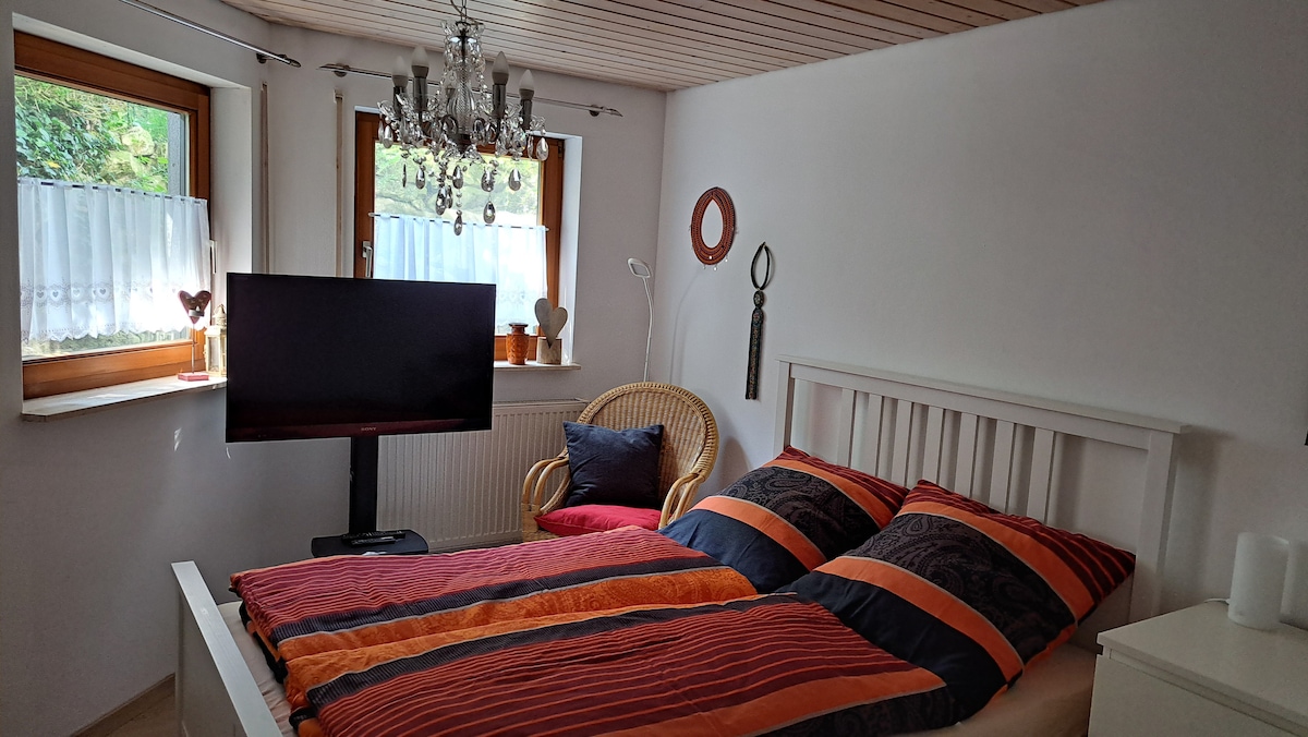A well-lit room is equipped with a spacious bed featuring orange and navy striped bedding. A flat-screen TV is positioned opposite the bed, and a rattan chair with a navy cushion is located beside a window, where natural light filters through sheer white curtains.