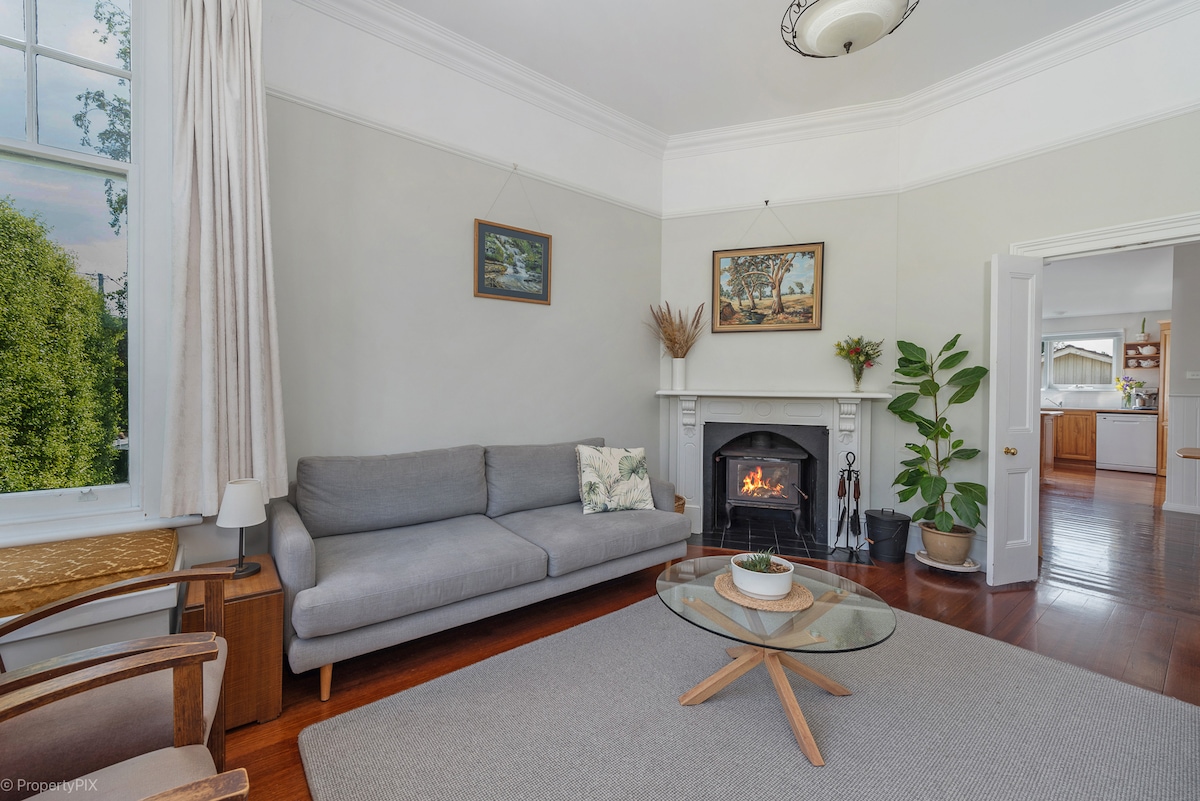 A cozy lounge area features a light gray sofa positioned in front of a fireplace, with a round wooden coffee table in the center. Natural light floods in through large windows, highlighting the wooden flooring and wall-mounted art.