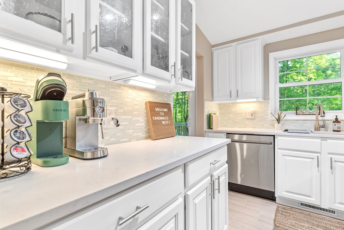 The kitchen features modern cabinetry with a clean white finish and sleek countertops. A gourmet coffee station is displayed, neatly organized with a coffee pod holder and various brewing options. Natural light streams through a window, illuminating the space and highlighting a serene outdoor view.