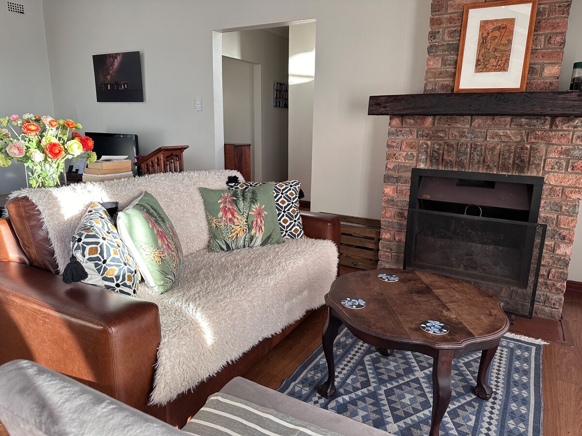 A warm living area showcases a brown leather sofa adorned with patterned cushions and a cozy throw. A wooden coffee table sits on a patterned rug in front of a brick fireplace, adding to the inviting ambiance of the space.