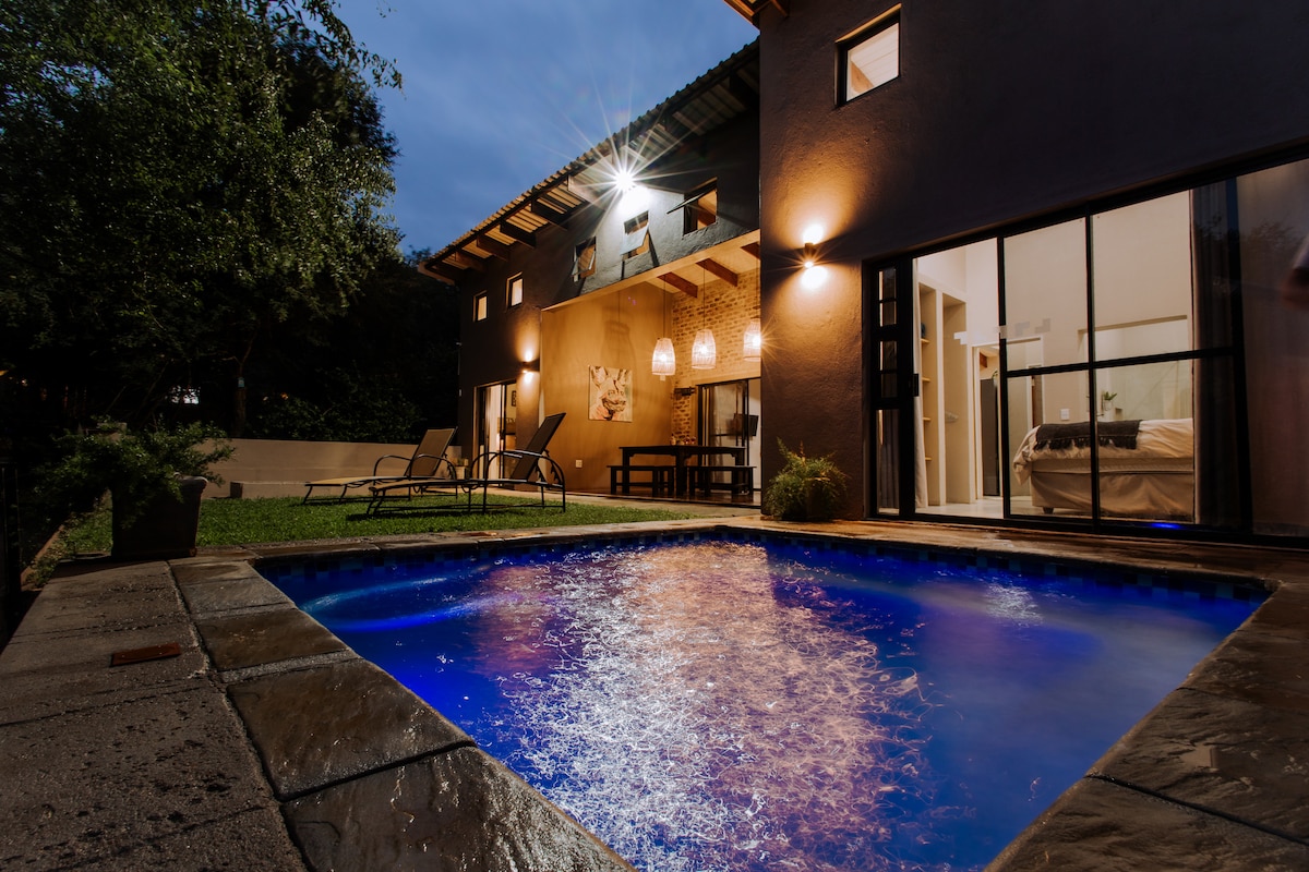 A tranquil pool area is illuminated with soft blue lighting, creating a serene ambiance. The house is framed by dark exterior walls and features glass doors that open to the interior. Lounge chairs are positioned nearby, surrounded by greenery and evening light.