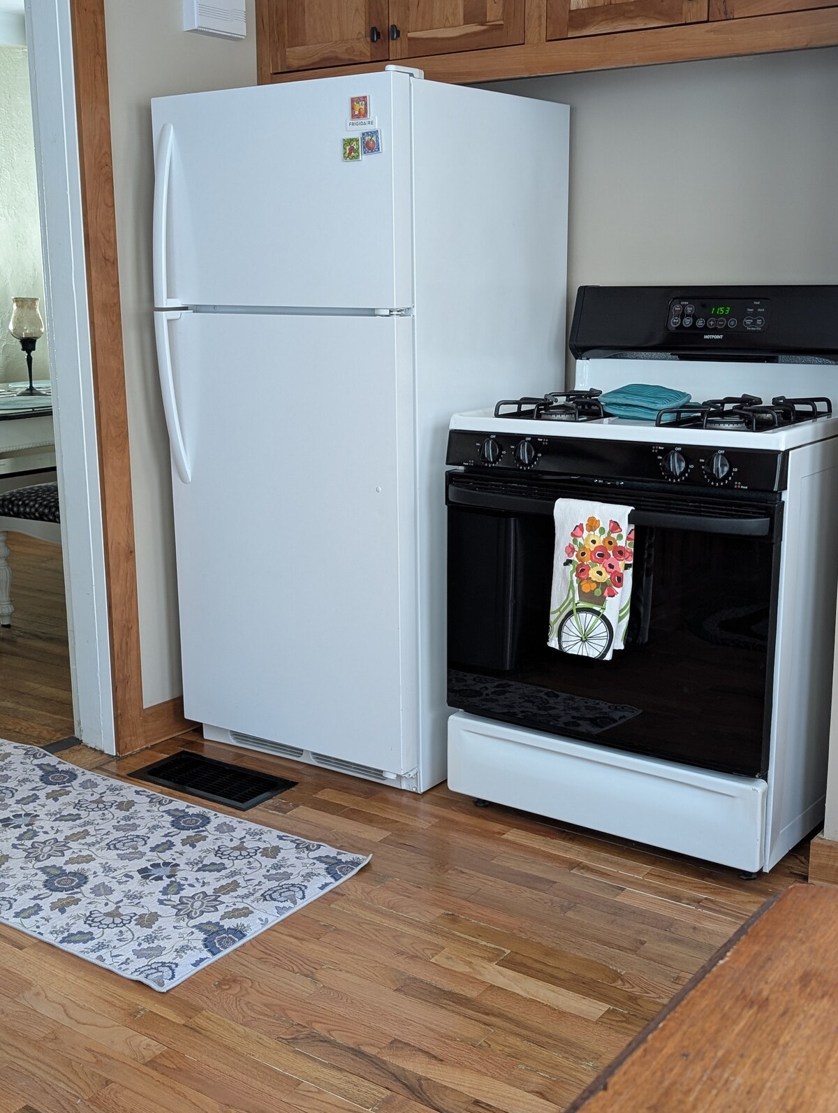 A clean and functional kitchen space includes a white refrigerator and a black gas stove. A colorful towel hangs on the stove, and a patterned rug lies on the hardwood floor, offering comfort underfoot.