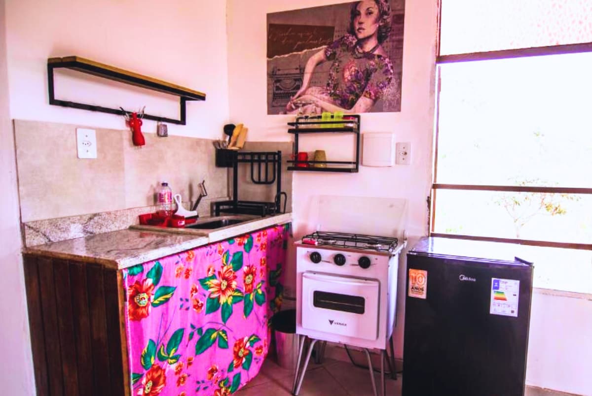 A compact kitchen area is presented, featuring a gas stove, refrigerator, and essential cooking utensils. Colorful floral fabric drapes the base of the countertop, while a wall-mounted shelf offers additional storage. Natural light filters in through a large window, enhancing the space's functionality.