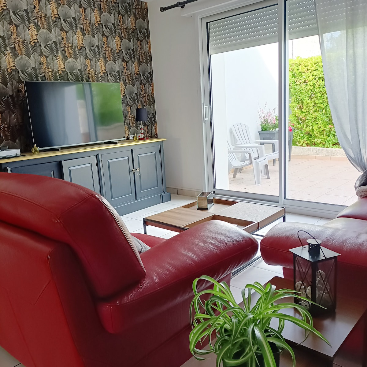 A living area is visible, featuring red leather seating arranged around a coffee table. A flat-screen television is mounted on a grey cabinet, while large glass doors open to a terrace with outdoor chairs. Natural light fills the space, highlighted by tropical-patterned wallpaper.
