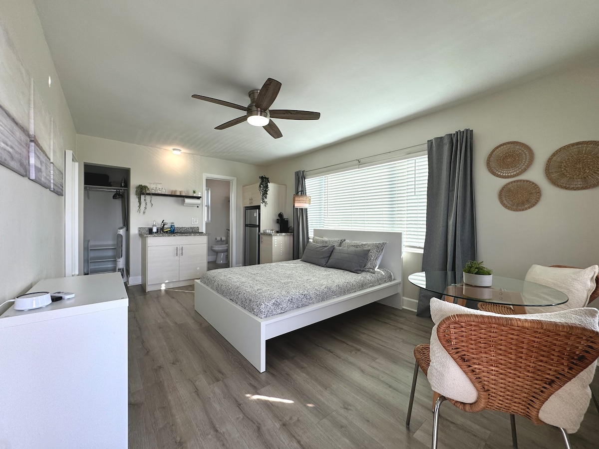 The studio apartment features a spacious bed with light linens, complemented by natural light from the window. A small dining area with two chairs is present, alongside a kitchenette area that includes a refrigerator and microwave. A ceiling fan adds to the room's comfort.