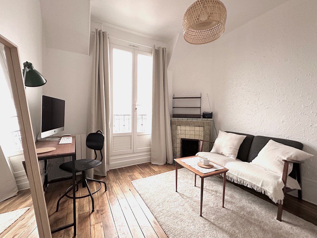 A bright living area features a comfortable two-seat sofa draped with a cozy blanket. A stylish desk with an iMac is positioned beside a chair. Natural light floods through the large windows, complemented by light-colored curtains, while a decorative fireplace adds a charming touch.