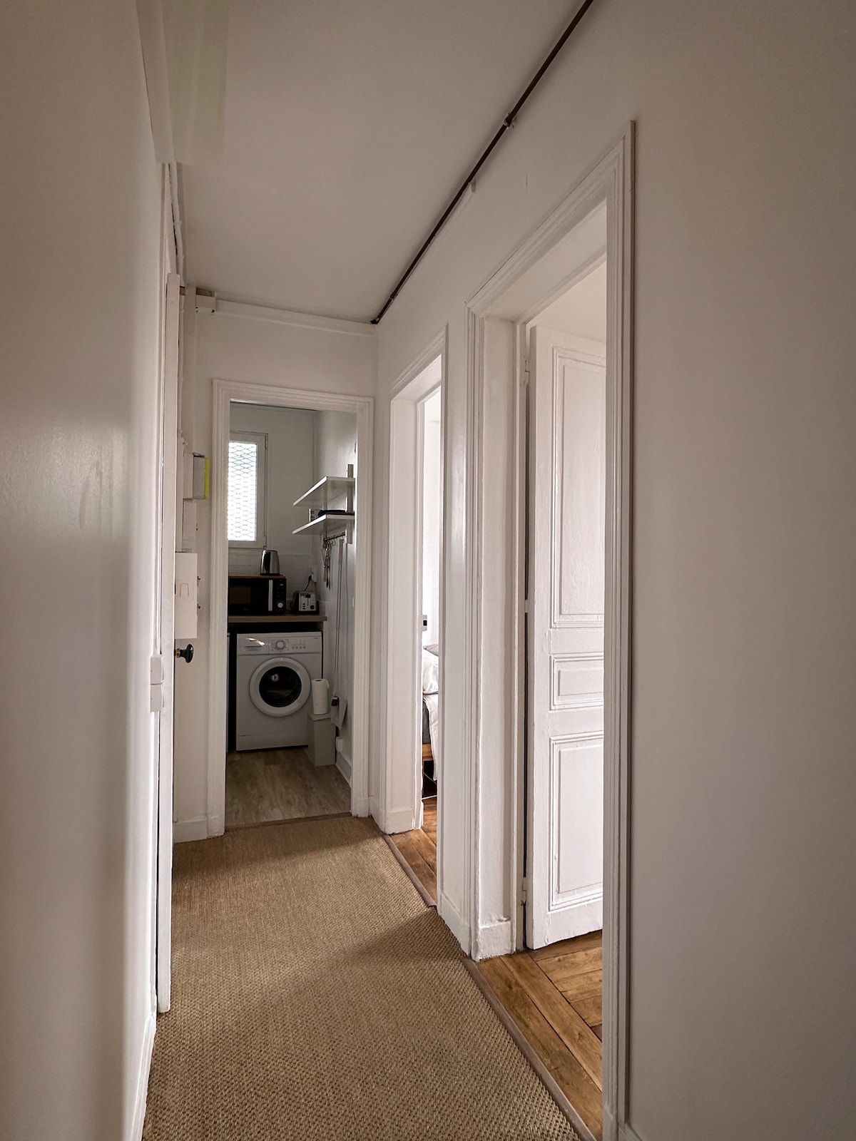 A well-lit hallway extends into the distance, featuring white walls and a natural fiber rug. Doorways lead to different rooms, including a laundry area equipped with a washing machine and shelves. The warm wood flooring adds a welcoming touch to the space.