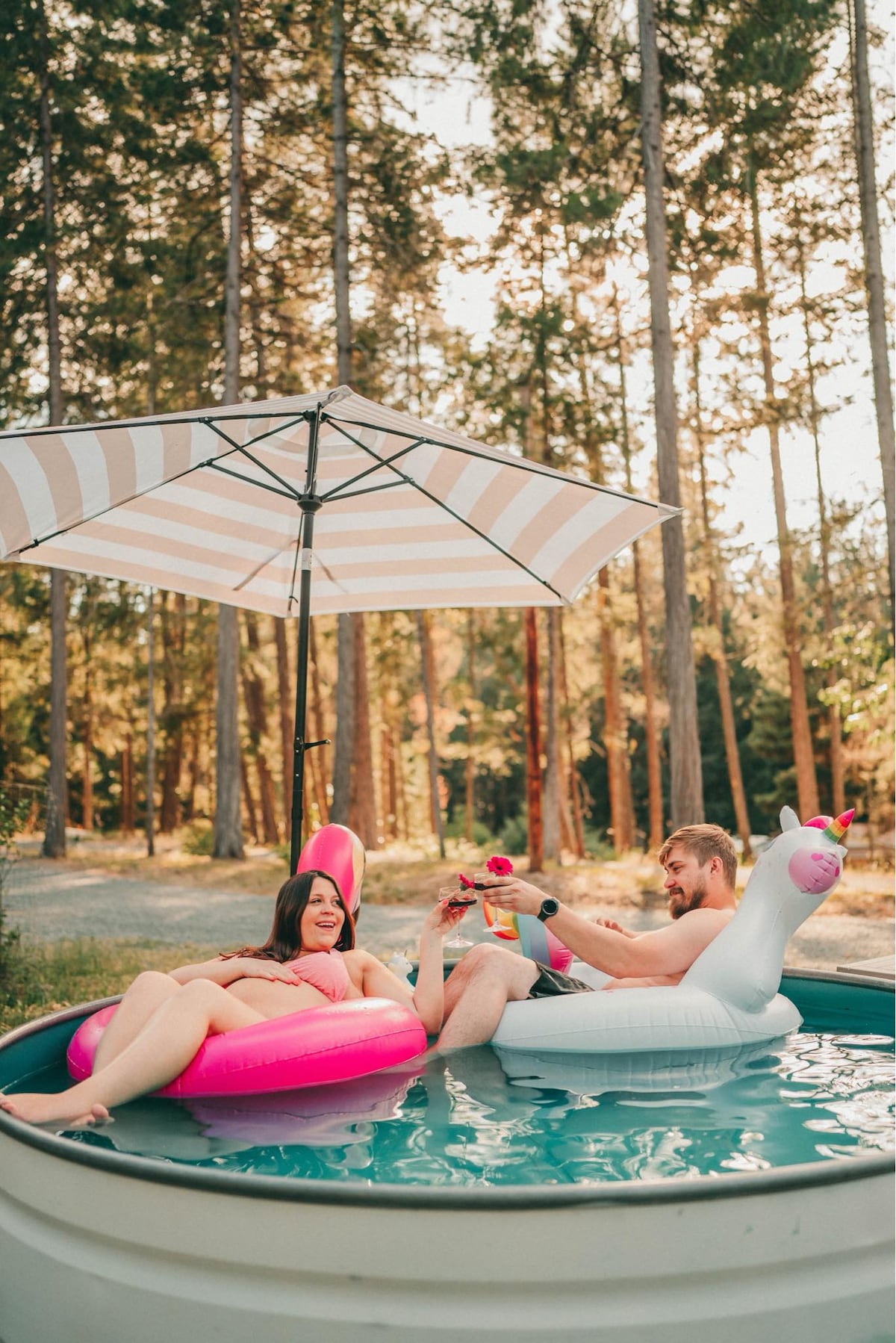 A small stock tank pool is surrounded by tall trees, offering a serene outdoor setting. Inflatable pool floats are visible, including a flamingo and a unicorn. A shaded area provided by a striped umbrella enhances the relaxing atmosphere.