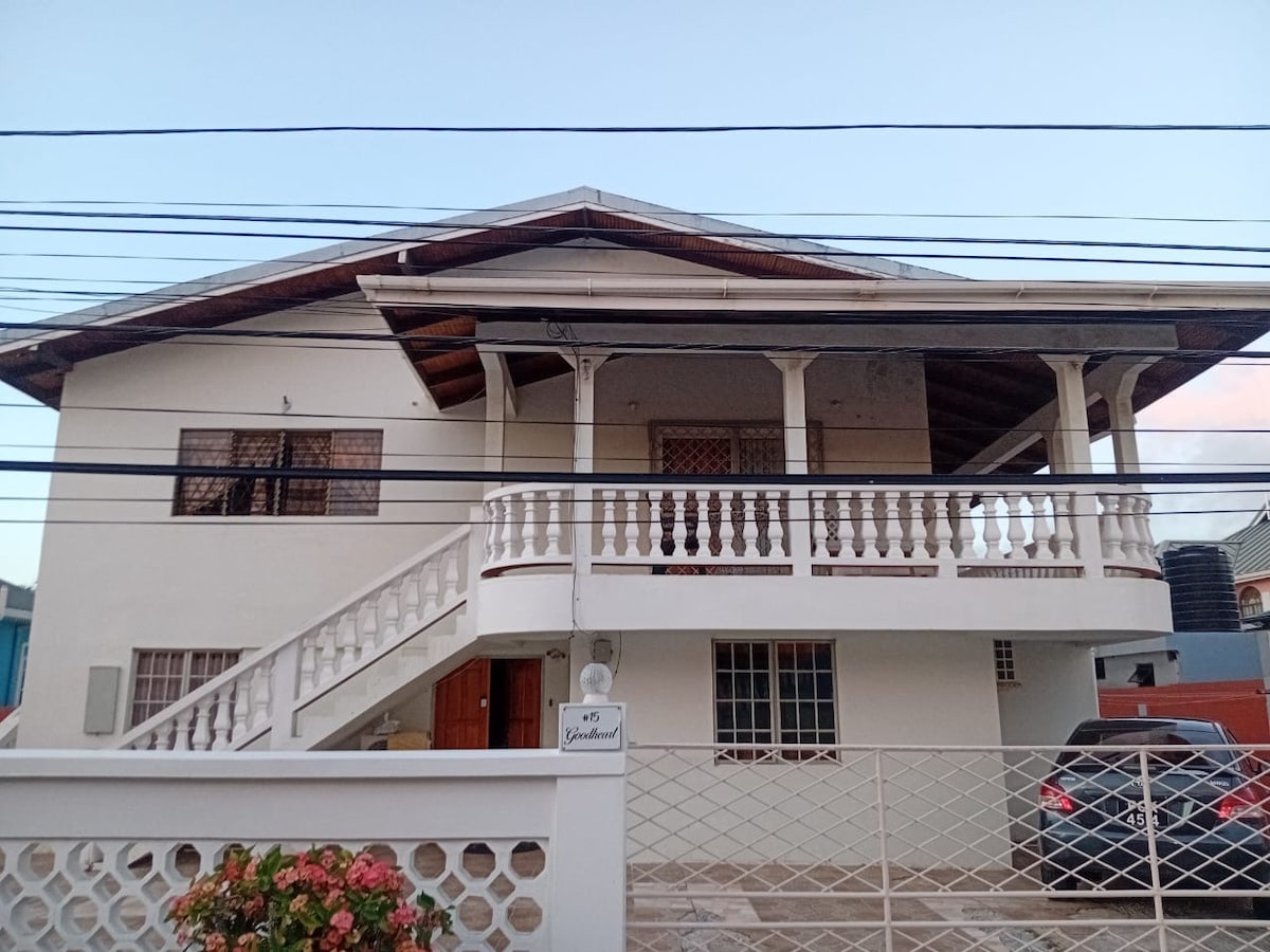 A two-story guest house is presented, featuring a spacious front porch supported by decorative columns. A staircase leads to the upper level, while the ground floor is accessible. The property is surrounded by a fence, with a flowering shrub visible in the foreground.
