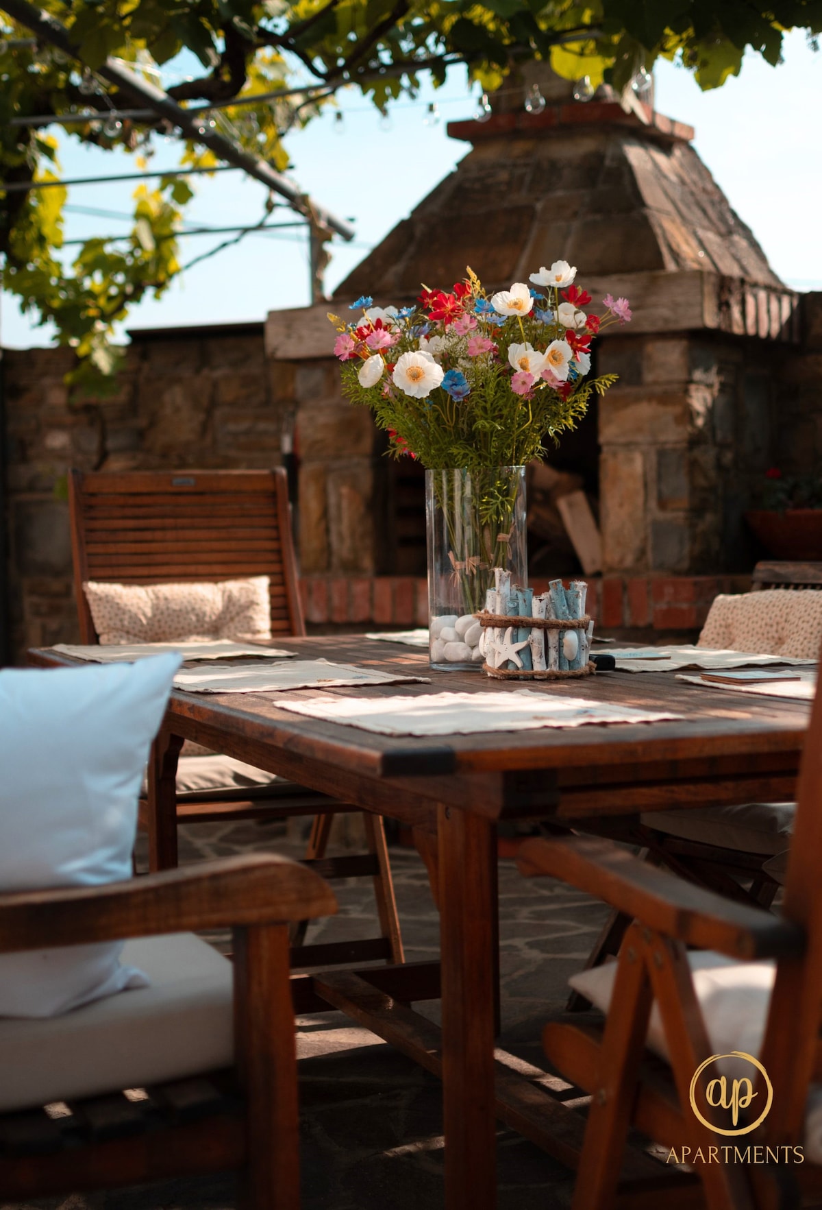 A dining table is set outside, surrounded by wooden chairs. A large vase filled with fresh, colorful flowers sits at the center, adding a natural touch. In the background, a traditional stone barbecue is partially visible, framed by leafy vines.