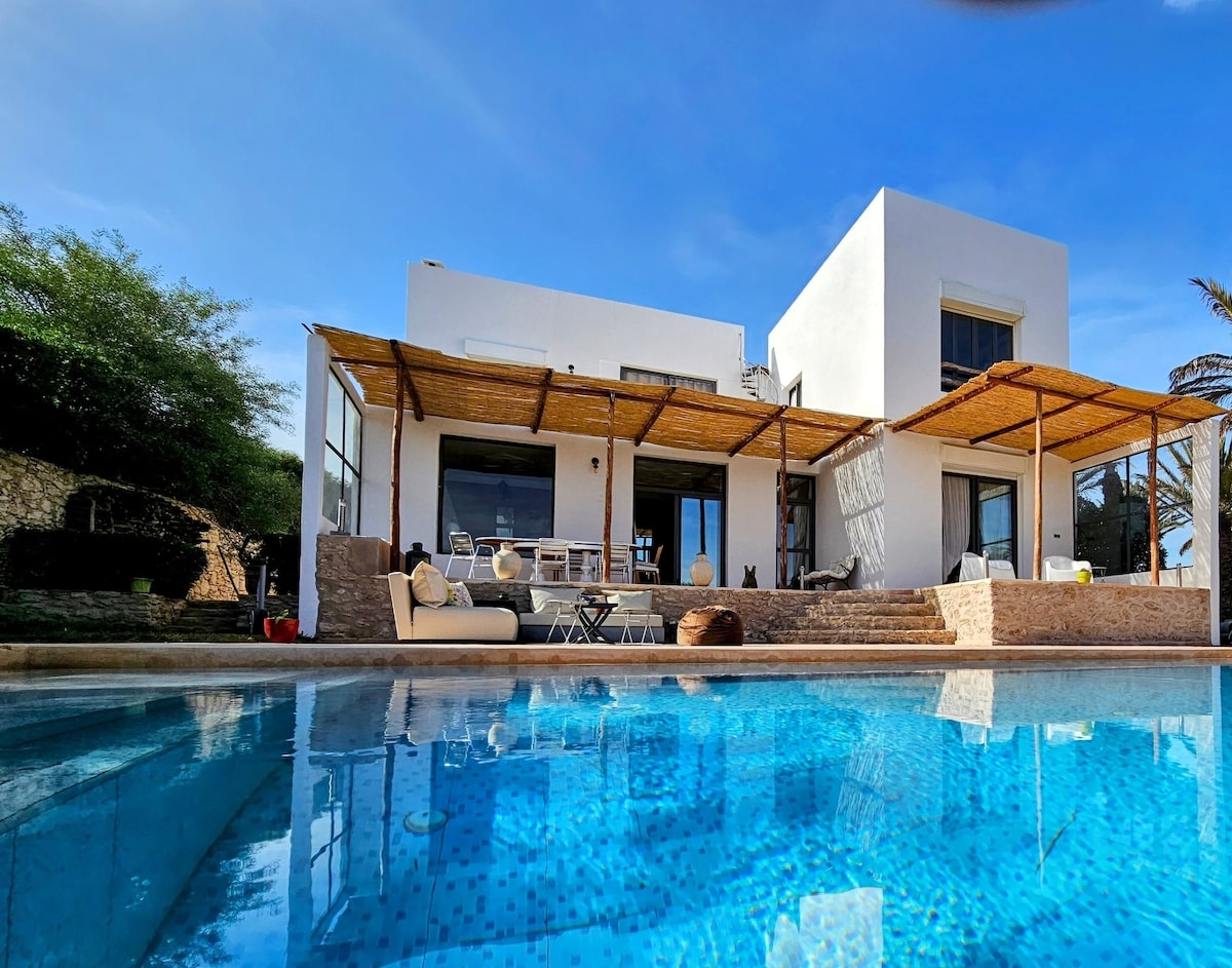 A modern villa features a spacious terrace shaded by bamboo, leading to a clear swimming pool. Large glass doors provide access to the interior, while multiple outdoor seating areas invite relaxation. The surrounding garden is adorned with greenery, creating a tranquil atmosphere.
