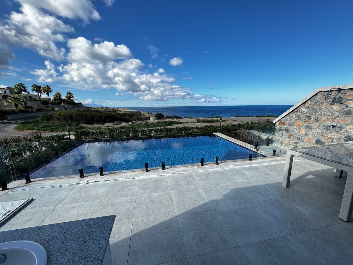 A large swimming pool sits surrounded by a spacious patio, with the sea visible in the distance. The sky is bright with scattered clouds, and coastal vegetation is present along the perimeter. The stone structure of the building is partially visible in the image.