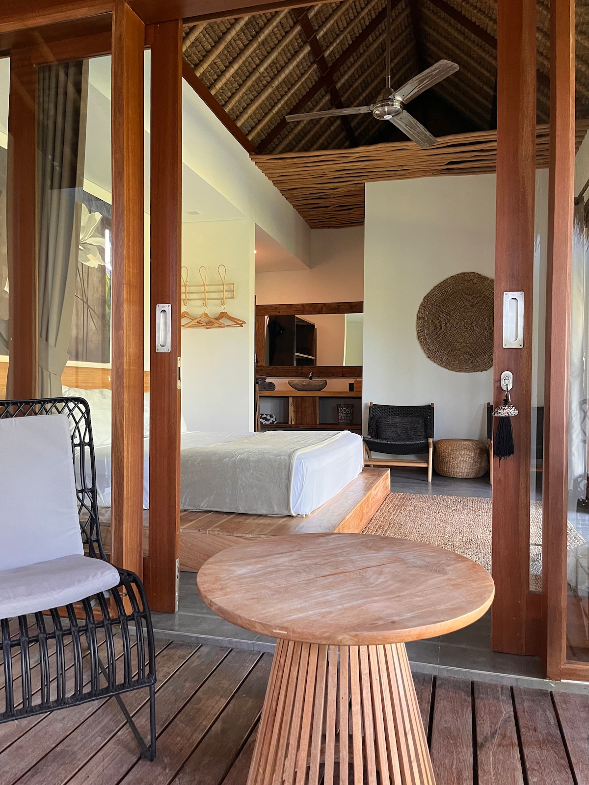 An inviting outdoor space features a woven chair paired with a round wooden table. Large glass doors provide a view into a well-lit interior with a spacious bed and natural elements. The thatched ceiling adds texture, while a decorative wall piece completes the room.