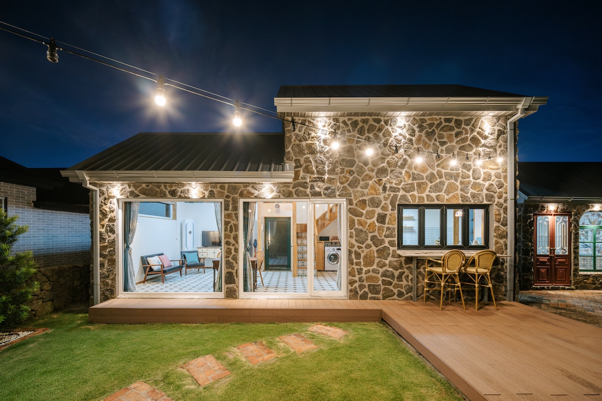A charming stone cottage is illuminated by exterior lights against a night sky. Large glass doors invite natural light into the interior, which reveals an open layout. A wooden deck extends into a grassy area, enhancing the serene outdoor space.