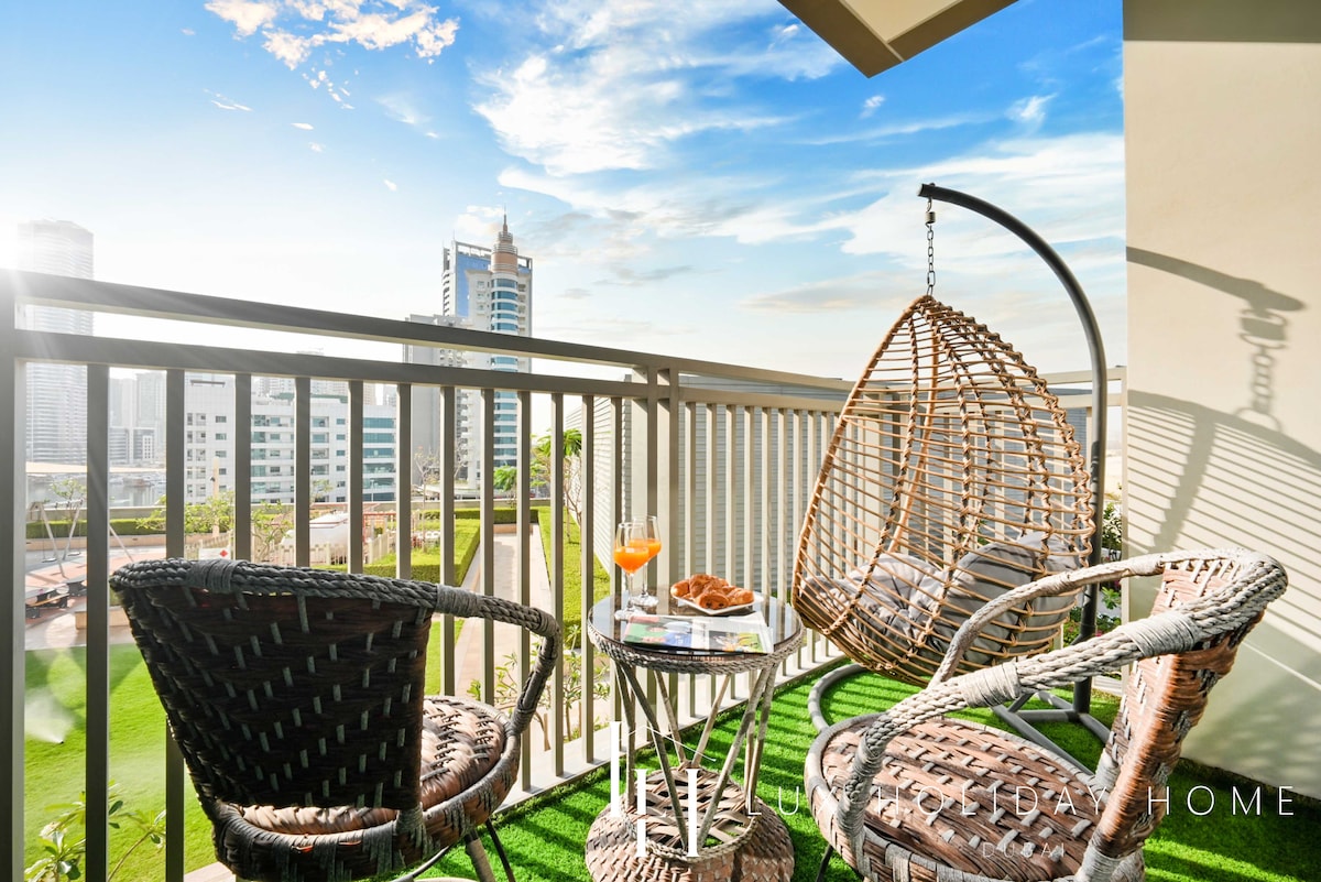 A serene private terrace features two woven chairs beside a round table. A bright orange drink and a plate of pastries are placed on the table. The view includes green landscaped areas and modern buildings under a clear blue sky.