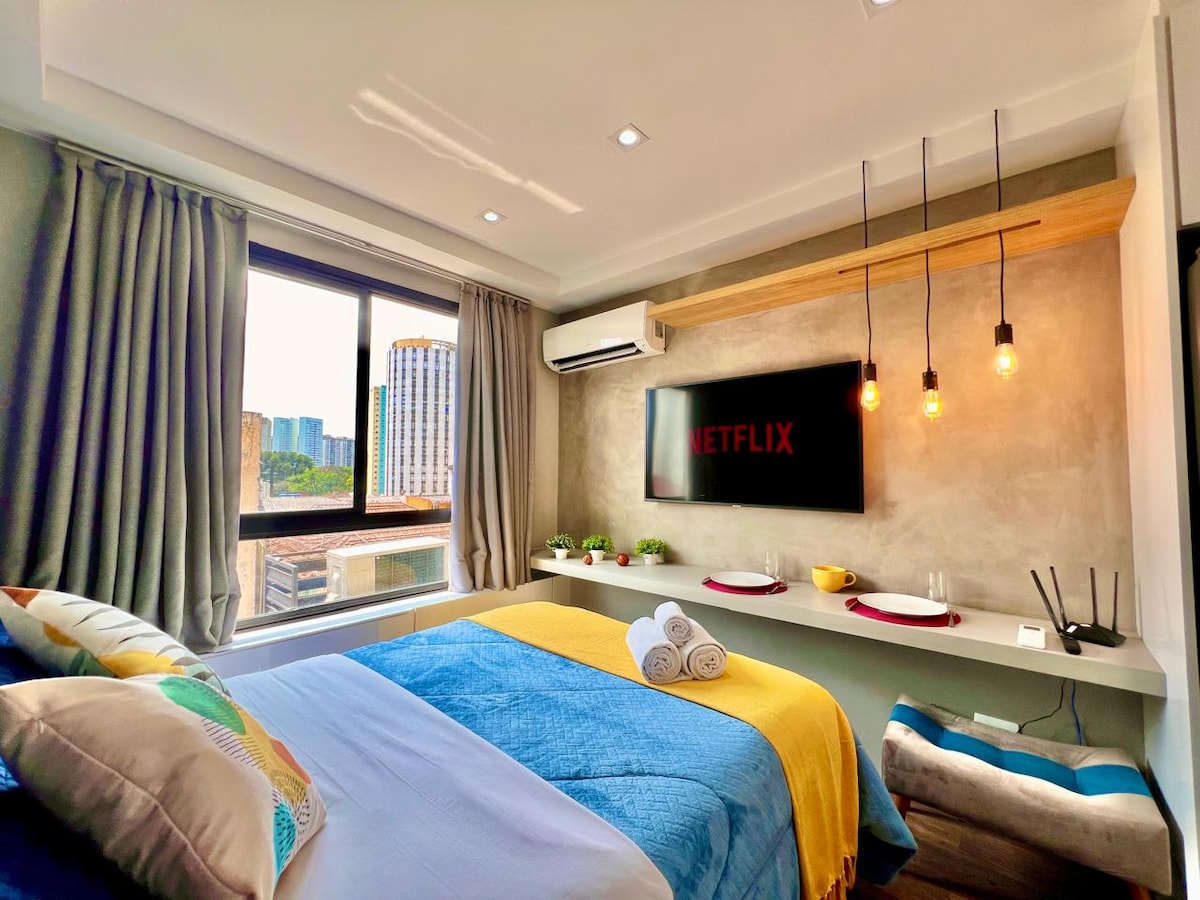 A cozy sleeping area features a spacious bed with colorful blankets and decorative pillows. A Smart TV mounted on the wall is accompanied by modern lighting fixtures. Large windows provide natural light and a view of the city skyline, enhancing the inviting atmosphere.