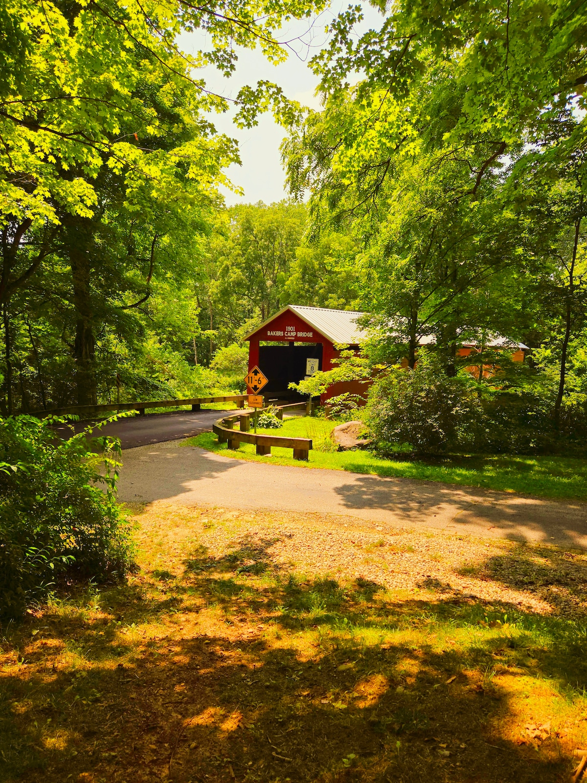A red covered bridge is framed by lush green trees, with a gravel path leading toward it. Sunlight filters through the foliage, creating a warm and inviting ambiance. The surrounding area is dotted with bushes and a manicured lawn.