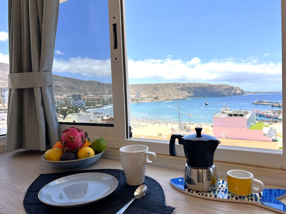 A breakfast setting is displayed by a window, showcasing a view of the beach and coastline. A variety of fruits and a coffee pot are arranged on a plate. The calm blue sea and distant mountains are visible in the background, illuminated by natural light.