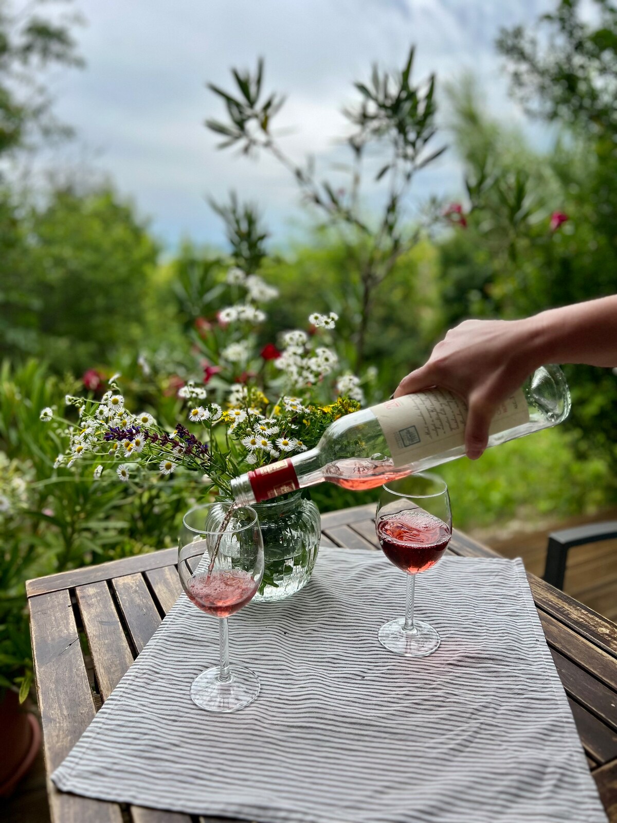 A hand is shown pouring wine into a glass on a table set with a striped tablecloth. Two glasses filled with pink wine are positioned next to a vase containing fresh flowers. Lush greenery is visible in the background.