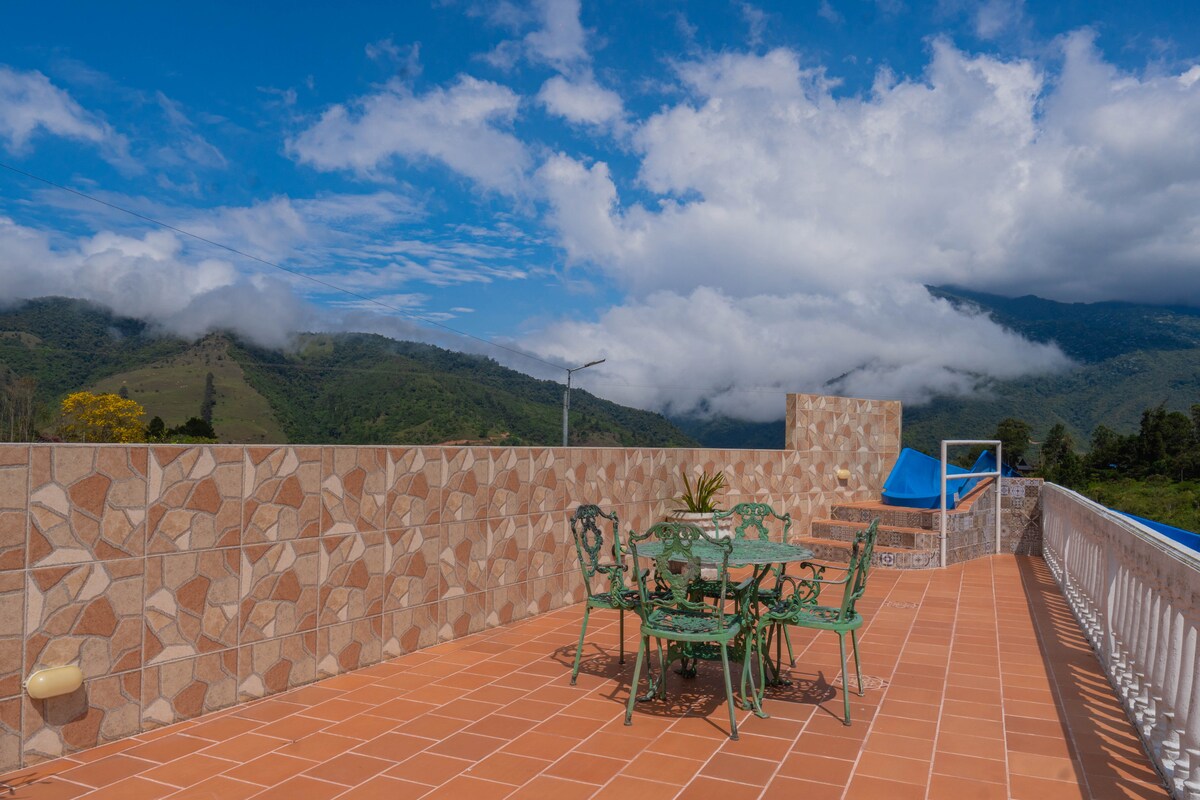 A spacious rooftop terrace features a tiled floor and green metal furniture, including a table and chairs. Surrounding natural landscapes and mountains are visible in the distance, with clouds adding texture to the sky.
