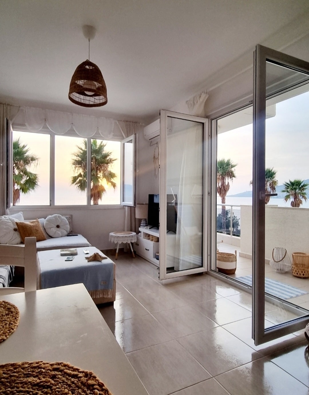 A modern living area features large glass doors that open to a balcony, allowing natural light to fill the space. The room includes a light-colored couch, a small coffee table, and a television. Potted palm trees are visible outside, complementing the coastal scenery.