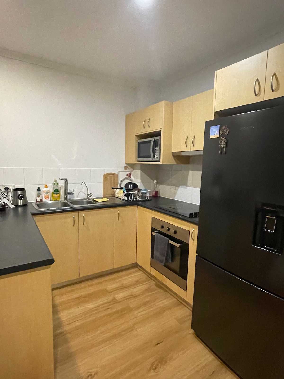 The kitchen features light wood cabinetry paired with black countertops, equipped with essential appliances including a microwave, stove, and refrigerator. Sink and countertop space are available for meal preparation, with bottles and utensils on the shelves, providing a functional cooking environment.
