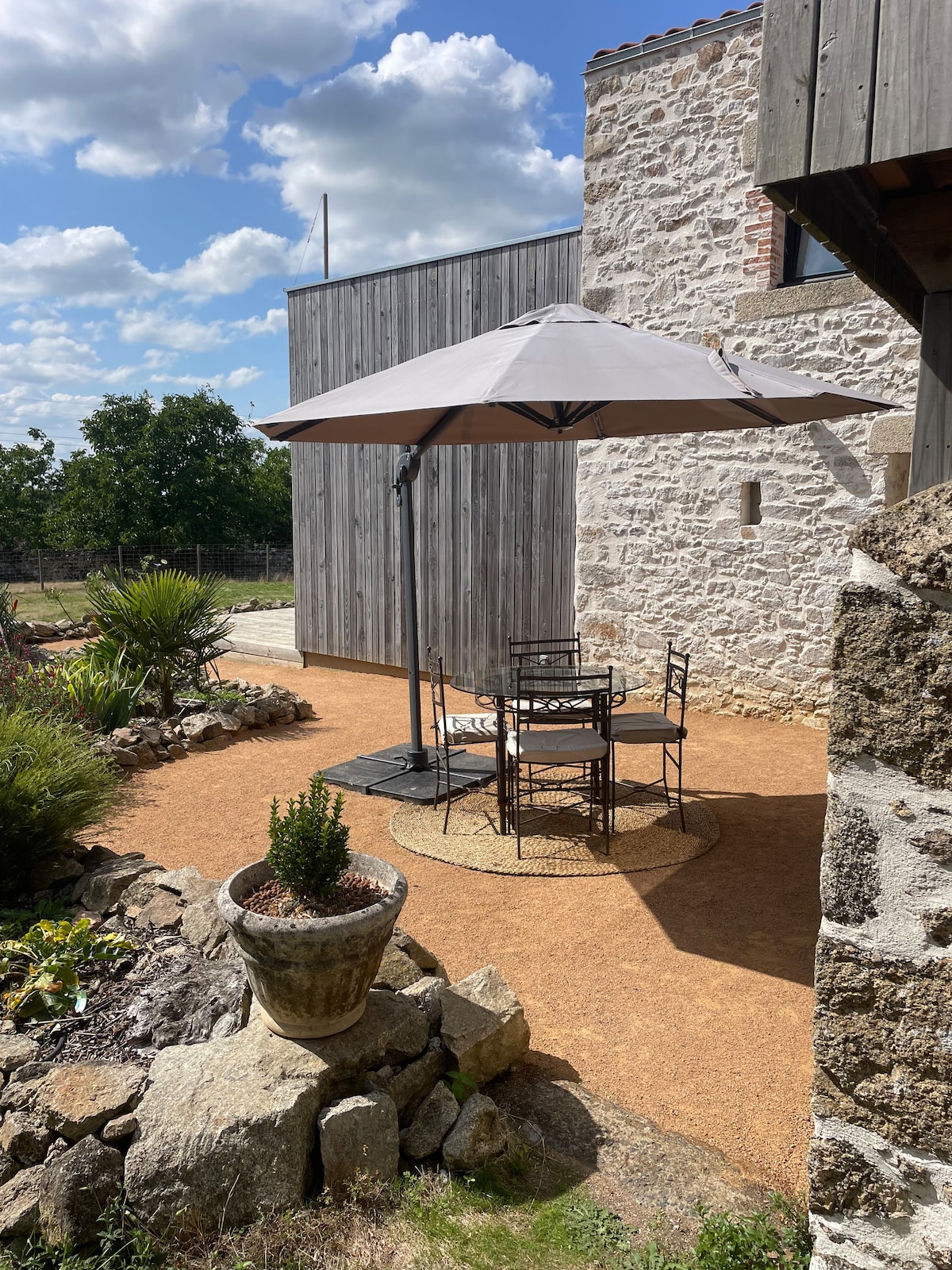 A private terrace is shown, featuring a round table with four chairs beneath a large umbrella. Surrounding plants add greenery, while the rustic stone wall of the building provides a textured backdrop. The path is made of packed earth, enhancing the outdoor ambiance.