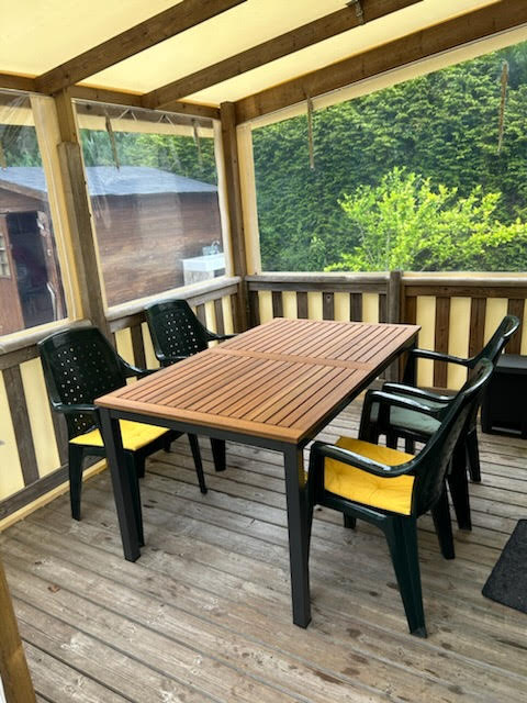 A covered terrace features a wooden table surrounded by four chairs with yellow cushions. The space is enclosed by clear panels, allowing natural light to filter in while providing views of the lush greenery outside.