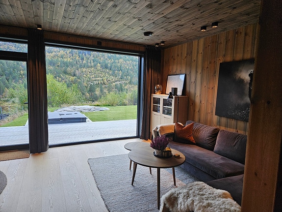A spacious living area features a dark sofa and a round coffee table atop a textured rug. Large glass doors open to a view of the surrounding nature and outdoor space, bringing natural light into the room. Wooden walls create a warm and cozy ambiance.