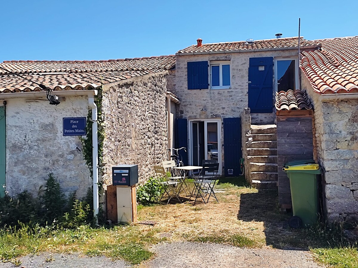 A stone house with blue shutters is depicted, featuring a staircase leading to the upper area. A small outdoor seating arrangement is visible with a table and two chairs. Lush greenery and a green trash bin are also present, under a clear blue sky.