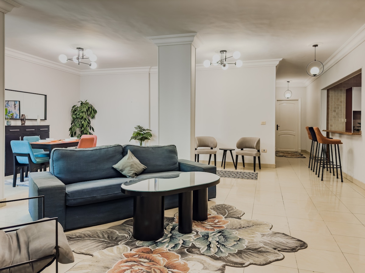 The spacious living room showcases a comfortable couch paired with a circular coffee table, complemented by a decorative area rug featuring floral patterns. A dining table with colorful chairs is visible in the background, along with additional seating at a kitchen bar.