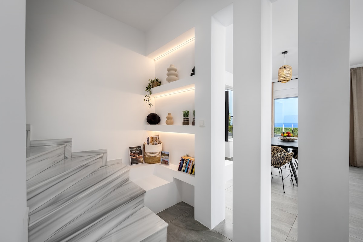 A bright and airy staircase with modern design features marble steps leading to an open space. Soft lighting accents minimalist shelving, displaying decorative items and a selection of books. A seamless view of the sea can be glimpsed through large windows in the background.