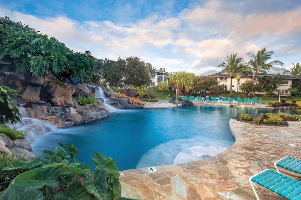 An expansive swimming pool is surrounded by lush tropical greenery and natural rock formations, including a small waterfall. Poolside loungers provide seating options, while palm trees sway gently in the background, contributing to a relaxing outdoor atmosphere.