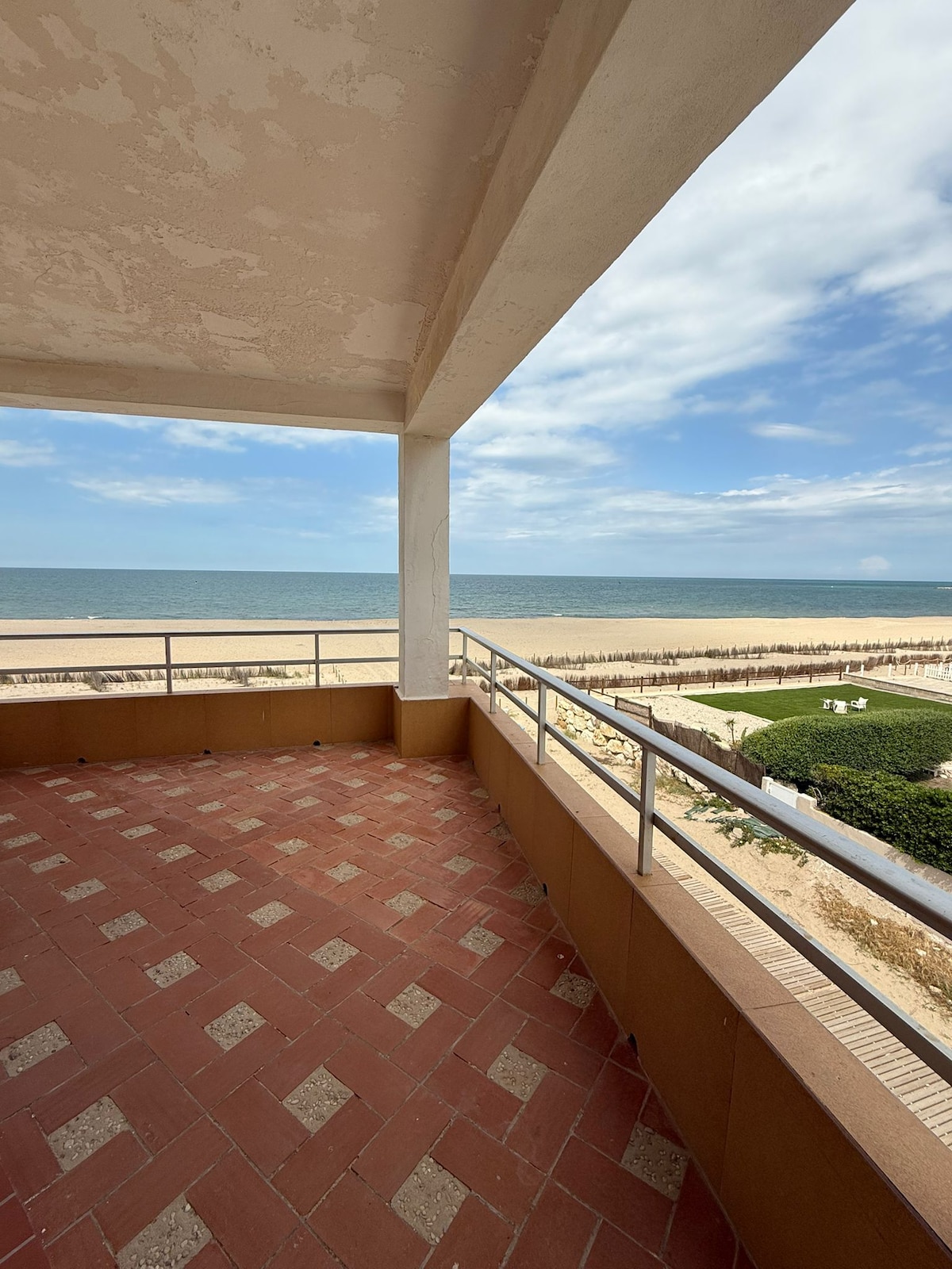 A spacious terrace offers a sweeping view of the beach and ocean. A railing surrounds the area, and the floor features a tiled pattern. The sky is partly cloudy, enhancing the coastal ambiance, while the sandy beach stretches into the distance.