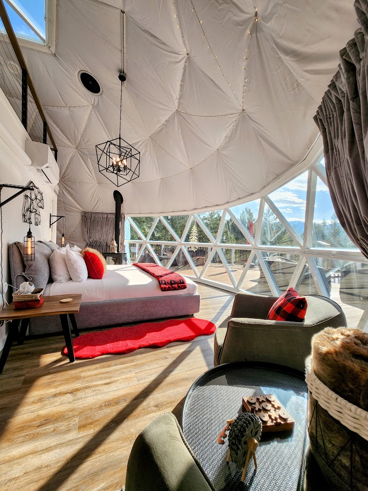 The interior of the dome features a spacious seating area with a plush bed adorned in neutral tones. Geometric windows allow natural light to flood in, highlighting wooden flooring. A cozy game table sits nearby, surrounded by comfortable chairs, creating a relaxed ambiance.