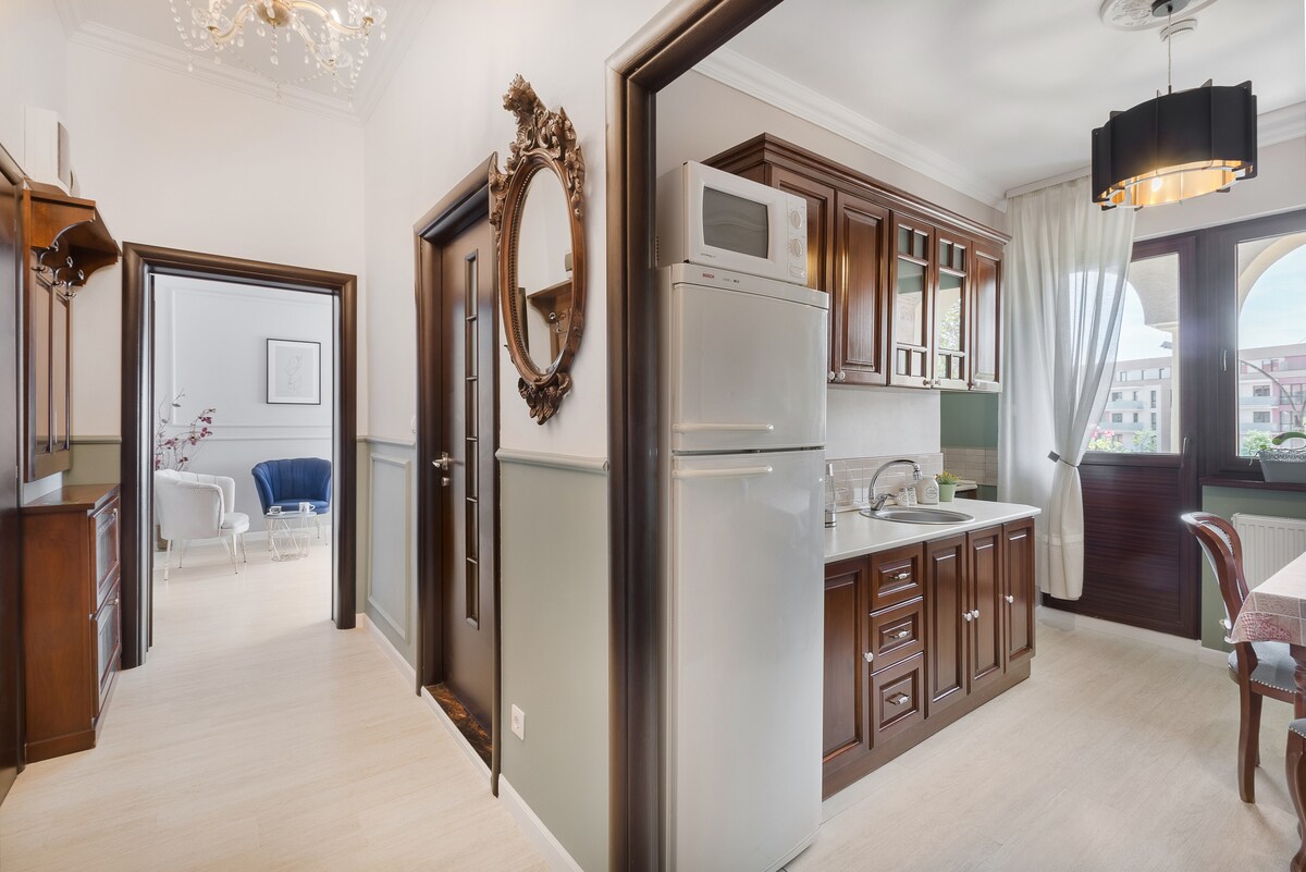 The kitchen area features dark wooden cabinets and modern appliances, including a refrigerator and microwave. Adjacent, a dining table is set for two. Light filters in through large windows, illuminating the space. The doorway leads into a brightly decorated living area.