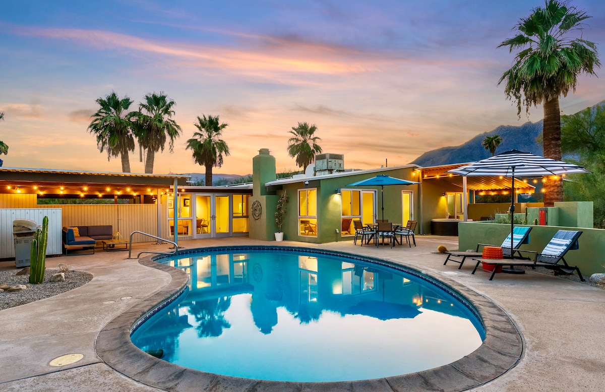 A serene pool area is framed by palm trees, with a vibrant sunset reflecting on the water's surface. Lounge chairs and an umbrella provide spacious seating options around the pool, while the house features large windows and a light, inviting exterior.