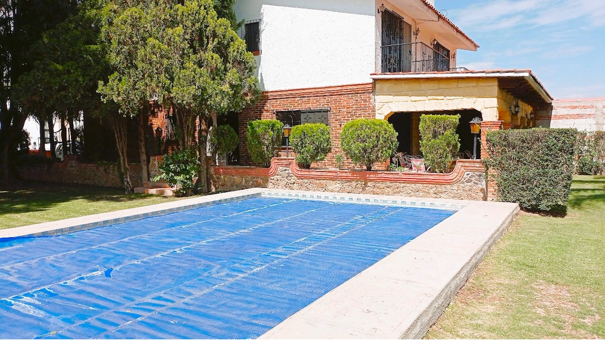 A spacious outdoor area features a covered pool with a safety cover, surrounded by well-maintained greenery and shrubs. The house is positioned in the background with a charming patio area visible. The setting provides an inviting environment for relaxation and gatherings.