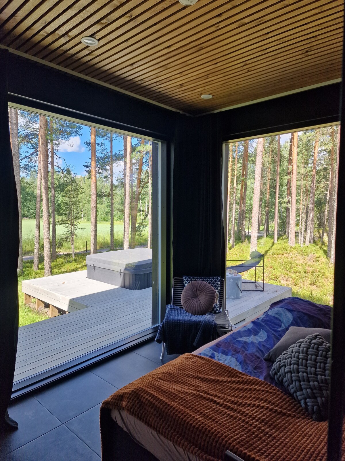 A cozy sleeping area features large glass windows with views of the surrounding forest. A double bed is dressed in colorful bedding, complemented by decorative pillows. A comfortable chair is positioned nearby, inviting relaxation as sunlight filters through the large openings.