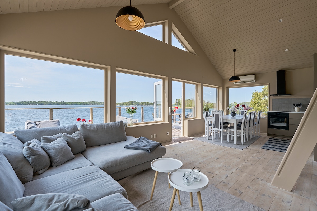A modern living area is highlighted by large windows offering views of the waterfront. A spacious gray sofa and minimalistic coffee tables are present. A dining area with white chairs is positioned beside the window. Natural light fills the room, creating a bright and open atmosphere.