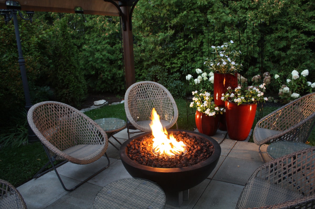 An inviting outdoor seating area features several wicker chairs arranged around a modern fire pit. Lush greenery surrounds the space, with vibrant red decorative pots filled with white flowers positioned nearby.