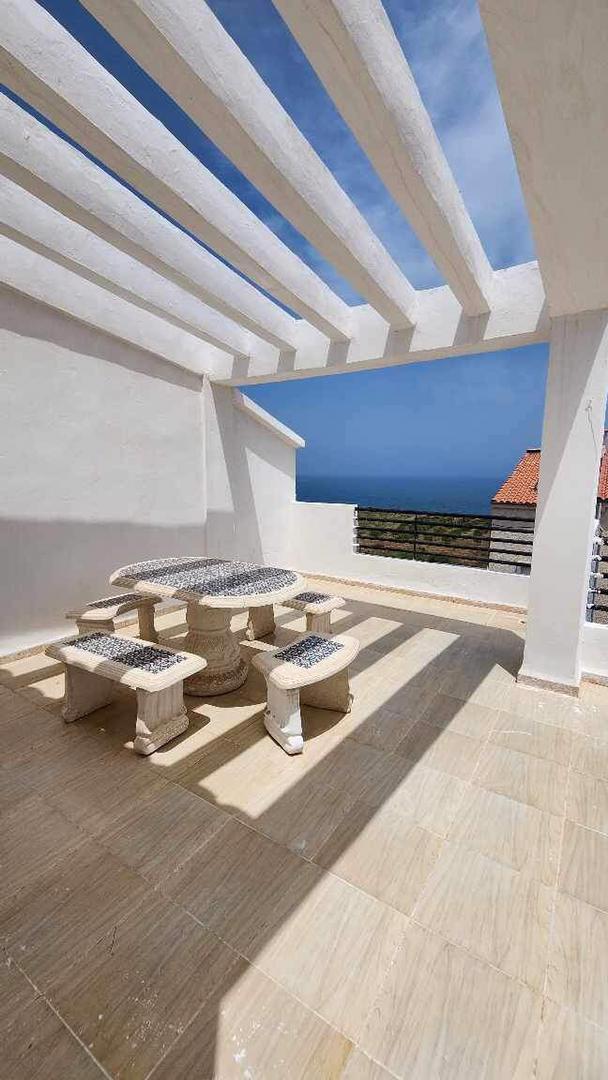 A spacious terrace is presented, featuring a circular stone table surrounded by four matching benches. The simplistic design is complemented by a gently sloped white ceiling, and the background reveals a clear view of the sea under a blue sky.