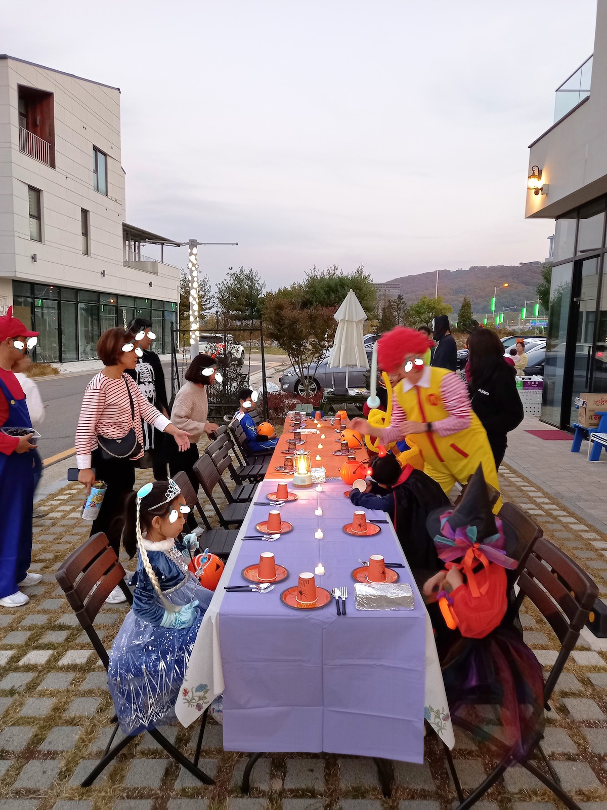 A festive outdoor dining setup is displayed, featuring a long table adorned with candles and orange decorations. Children in various costumes engage playfully at the table, while adults assist in the background. The setting is complemented by trees and buildings visible in the distance under an evening sky.