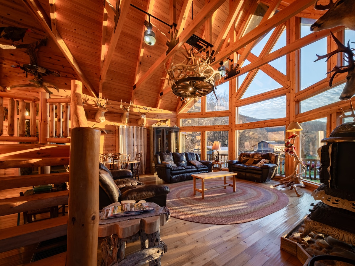 The spacious interior of a log cabin reveals a high, wood-beamed ceiling and multiple large windows that allow natural light to flood in. Comfortable seating arrangements include black leather sofas and a wooden coffee table, enhancing the inviting atmosphere. Rustic decor elements are visible throughout.